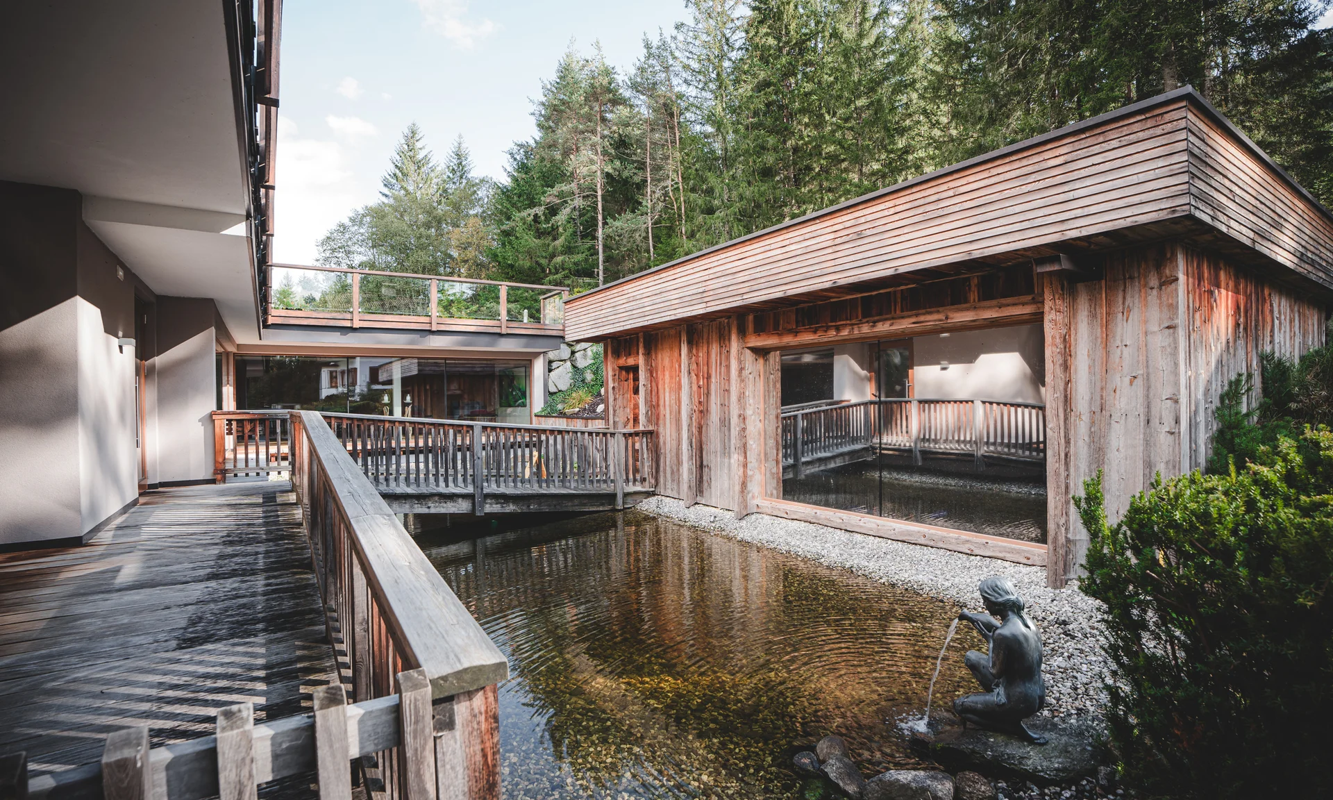 Entschleunigen und genießen in diesem Hotel nahe Bruneck Holzterrassen und Teich mit Brunnenfigur in einem Waldresort