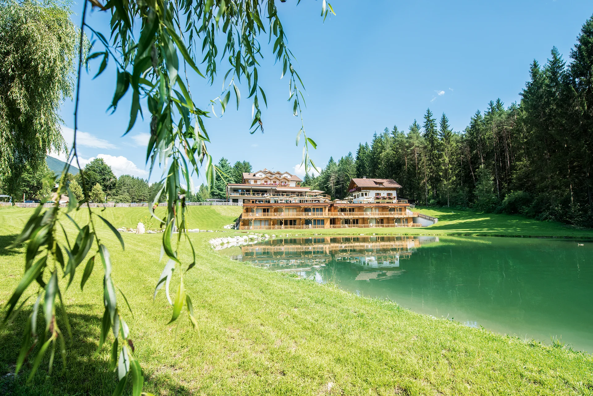 Entschleunigen und genießen in diesem Hotel nahe Bruneck Hotelgebäude am Seeufer mit grüner Wiese und Wald im Hintergrund