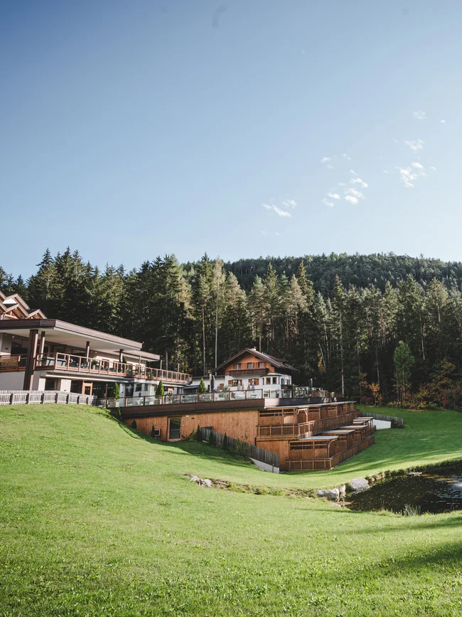 Entschleunigen und genießen in diesem Hotel nahe Bruneck Hotelgebäude mit Terrasse neben Teichen und Wald unter klarem Himmel