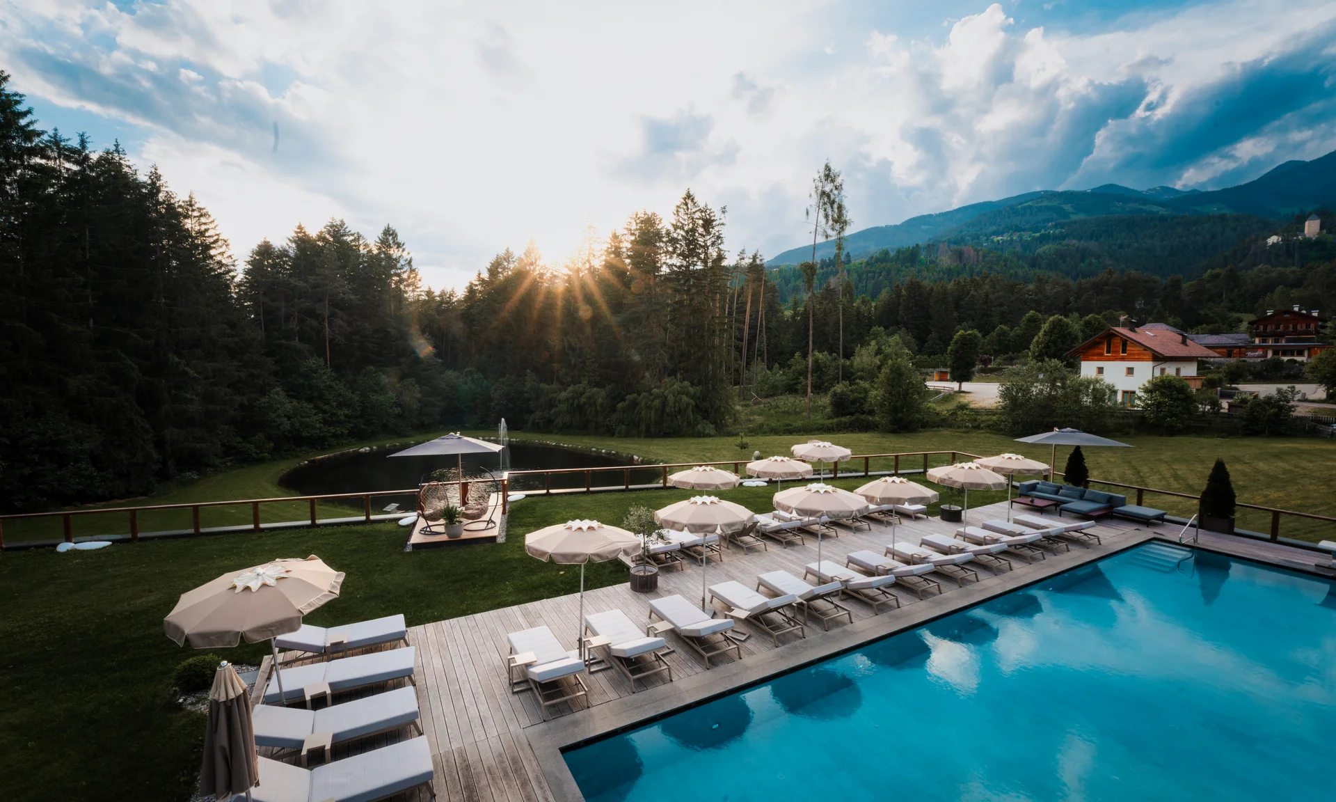 Entschleunigen und genießen in diesem Hotel nahe Bruneck Pool mit Liegestühlen und Sonnenschirmen vor Wald und Bergen bei Sonnenuntergang